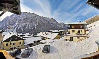 A winter landscape with snow-covered hills and houses. In the background, tall mountains stretch under a clear sky.