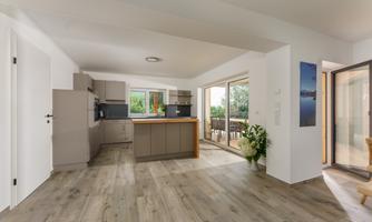 A modern kitchen with an open living area and large windows. The room has bright walls and an elegant wooden floor.