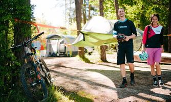 Two cyclists are on a campsite. In the background, tents and bicycles can be seen, surrounded by trees.