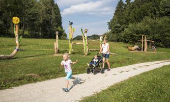 A mother is pushing a stroller on a path while a little girl happily runs ahead. In the background, there are artistic wooden sculptures and green meadows.