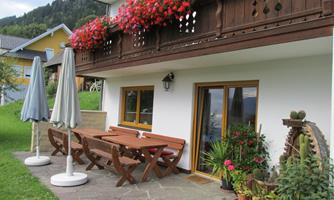 A terrace with a large wooden table and benches, surrounded by colorful flowers. In the background, there are parasols and a green garden.