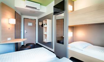 A modern hotel room with two single beds and a small table. The walls are painted in neutral colors and there is plenty of storage space.