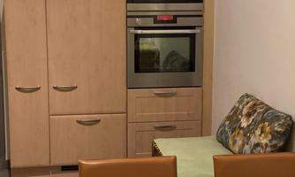 A modern kitchen with a wooden cabinet and a double oven. In the foreground, there are two chairs and a table with a tablecloth.