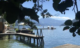 A quiet lake with boats and a wooden terrace. The landscape is surrounded by trees and mountains.
