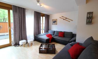 A cozy living room with a gray sofa and red cushions. Large windows let in plenty of light and offer a view of the outdoors.