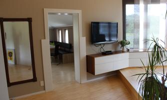 A modern living room with a light wooden floor and large windows. The room features a flat wall TV, a low table, and a large mirror.