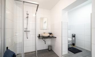 A modern bathroom with a shower and large white tiles. The washing area includes a sink and a mirror.
