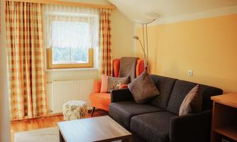 A cozy living room with a gray sofa and an orange armchair. Bright curtains and a table with a rug create an inviting atmosphere.