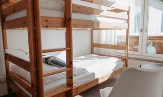 A simple bedroom with a wooden bunk bed and a white chair. There is plenty of daylight from the window.