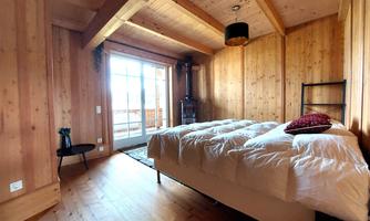 A cozy bedroom with wooden paneling and a large window. The bed is adorned with white bedcovers and a red pillow.