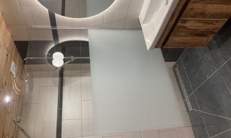A modern bathroom with a shower and a sink. The walls are light tiled, and there is a large, round mirror light.