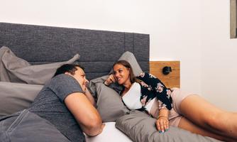 A couple lies relaxed in bed, smiling at each other. They seem to be having a conversation and enjoying their time together.