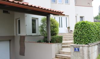 A bright house with an entrance area surrounded by a green garden. The stairs lead to a white door.