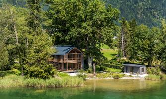 A beautiful wooden house on the shore of a clear, green lake, surrounded by trees. In the background, gentle hills can be seen.