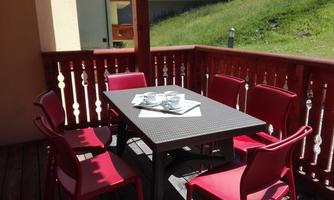 A cozy balcony with a table and six red chairs. There are glasses and a newspaper on the table.