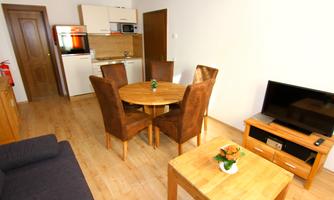 A modern living room with a round dining table and four chairs. The kitchen is open, and a television is placed on a wooden piece of furniture.