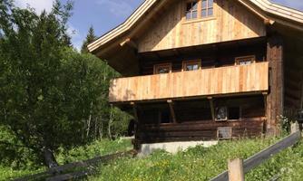 A cozy wooden house with a balcony, surrounded by green meadows and trees. The sky is clear and blue.