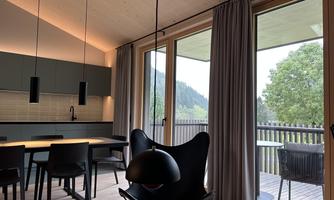 A modern apartment with large windows overlooking a green landscape. The space is bright and stylishly furnished with a dining area and a cozy armchair.