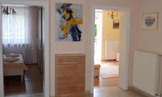 A bright hallway with a colorful carpet and a wooden cabinet. In the background, two rooms can be seen, accessible through open doors.