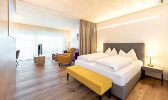 A modern hotel room with a large bed and yellow stools. Large windows provide plenty of daylight and a cozy atmosphere.