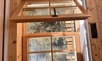 An open wooden window with a view of a wooded background. The sun is shining through the window and illuminating the room.