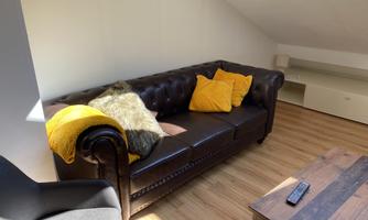 A cozy living room with a brown sofa and yellow cushions. The room has a wood-colored floor and plenty of natural light.