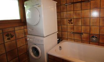 A bathroom with a washing machine and a dryer stacked on top of each other. In the background, there is a bathtub and tiled walls in warm colors.