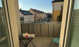 A small balcony with a table and two chairs. In the background, a quiet street with buildings and a clear sky can be seen.