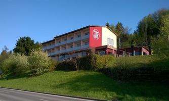 A modern building with several floors stands on a hill. It has a red façade and is surrounded by green nature.