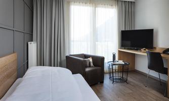 A modern hotel room with a single bed, a comfortable armchair, and a table. Large windows let in plenty of light and offer a view outside.