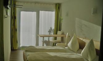 A bright bedroom with two beds and large windows. In the background, a table and a chair can be seen.