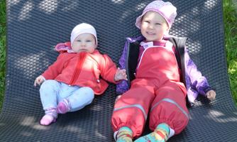 Two small children are lying on a mat outdoors. One child is wearing a red suit and the other a red jacket.