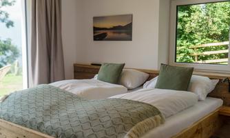 A cozy bedroom with a double bed and cushions in soft colors. Through the large window, you look out into the greenery.