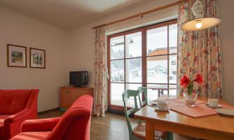 A cozy living room with red armchairs and a dining table. Large windows offer a view of the outdoor area.