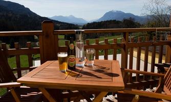 A cozy table on a porch with two chairs. In the background, mountains and a clear blue sky can be seen.