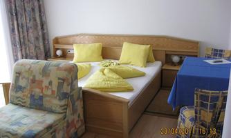 A cozy bedroom with a double bed, yellow pillows, and a seating area. A blue table stands next to the bed, and there are colorful curtains.