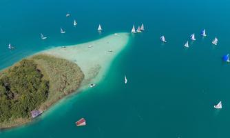 A picturesque coastal landscape with a small island and many sailboats on the turquoise water. The sky is clear and the beach is sandy and lush with greenery.