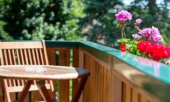 A cozy balcony with a wooden table and chairs. Colorful flowers are blooming nearby and the sun is shining.