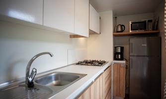 A modern kitchen with a sink, a stove, and a refrigerator. The furniture is made of light wood and the walls are kept simple.