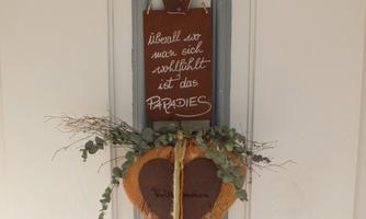 A heart-shaped welcome sign made of wood hangs on a door. Above it is a sign with the saying: "Wherever you feel comfortable, there is paradise."