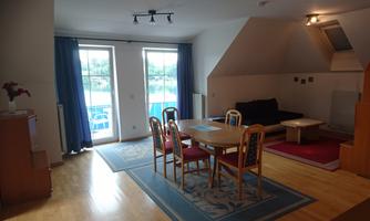 A bright living room with a dining table and chairs. Large windows offer a view of the greenery outside.