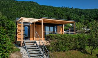A modern wooden house with large windows and a balcony, surrounded by green grass and bushes. In the background, gentle hills and a clear blue sky are visible.