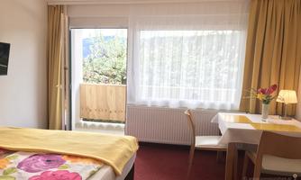 A bright room with a bed, a table, and chairs.  
The window leads to a balcony with a view of the greenery.