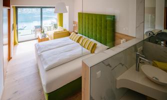 A modern bedroom with a green headboard and light colors. Large windows offer a view of the water and plenty of natural light.