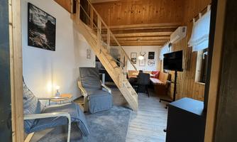 A cozy interior with paneled wooden walls and a staircase. On the left side, there are two chairs and a table, while on the right, a couch and a television can be seen.