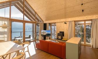 A modern living room with large windows and a view of the lake. There is a red sofa, a table, and a television.
