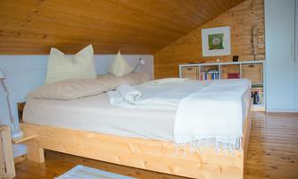 A cozy bedroom with a wooden bed and pillows. The walls are light, and there are bookshelves in the background.