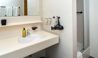 A modern bathroom with a bright sink and a large mirror. In the background, there is a shower and hygiene products on the shelf.