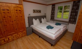 A cozy bedroom with two single beds and natural wooden furniture. The room has large windows that let in daylight.