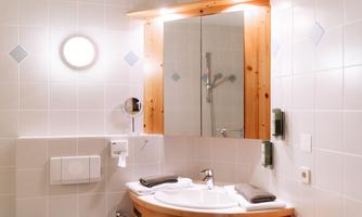 A modern bathroom with a round mirror and wooden paneling. The walls are light tiled and there is a shower in the background.